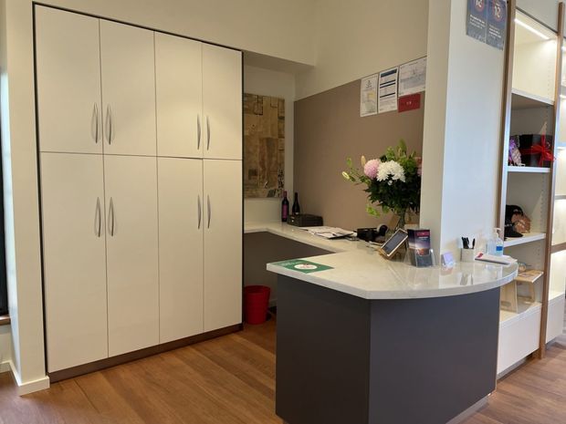 A compact reception or service counter area with a curved dark-front desk and pale stone-like countertop. Tall cream built-in cabinets with integrated handles line the left wall, while open shelving and storage occupy the right side. The room has warm timber flooring, muted taupe walls, work documents, stationery, and a floral arrangement on the counter. The space appears practical, tidy, and softly lit.