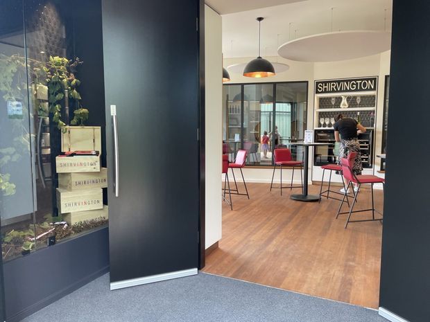 View through a dark-framed entrance into a stylish commercial tasting or retail space. The interior features warm timber flooring, black-framed glazed partitions, red and black bar stools and chairs, pendant lighting, a large oval acoustic ceiling panel, and a prominent SHIRVINGTON display wall with shelving. A planted display is visible beside the entrance.