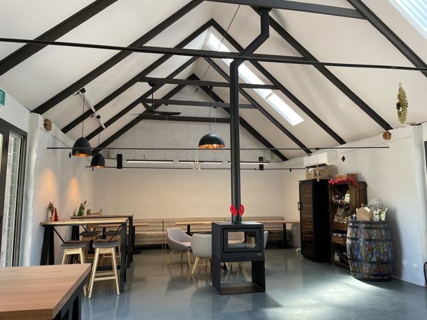 A bright, open-plan interior with a high pitched ceiling and prominent exposed black timber or steel trusses. White walls and ceiling contrast with a polished grey concrete floor. The space includes a dining area with timber tables and stools, pendant lighting, a long built-in bench, a central freestanding wood-burning stove, and a rustic barrel-style feature. Skylights bring in natural light, while dark structural framing creates an industrial-rustic character.