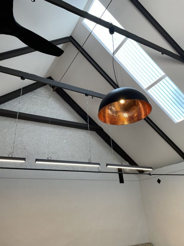 Upward-looking view of an interior with a high pitched ceiling, exposed dark structural beams, a large linear skylight, suspended linear LED lights, and a prominent black pendant light with a warm copper-toned interior. The walls are pale and minimally detailed, creating an industrial-contemporary character.