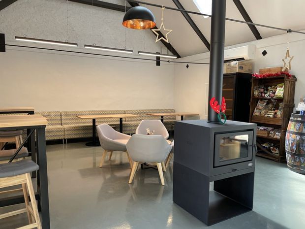 A modern, high-ceilinged communal seating area with polished concrete flooring, exposed dark structural beams, suspended linear lighting, and a black freestanding wood-burning stove. The room includes a long upholstered banquette, small round tables, contemporary upholstered chairs, bar-height seating, and open shelving with supplies. Festive star decorations and a few retail items are visible, suggesting a café, hospitality venue, or commercial lounge.
