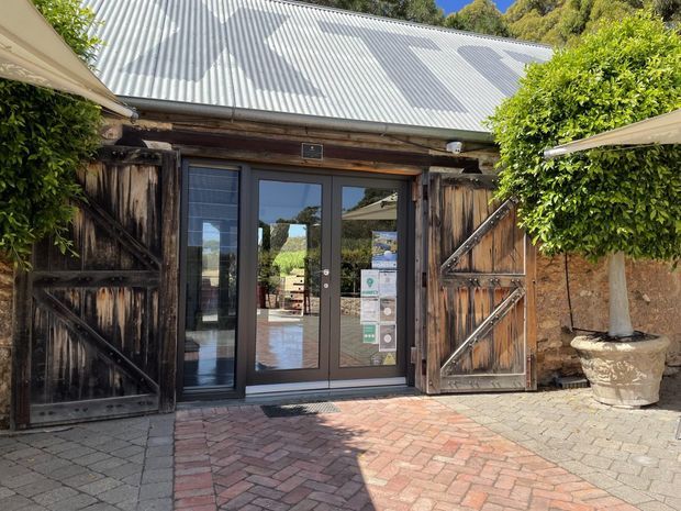 Exterior entrance to a rustic timber-clad building, featuring large weathered barn-style doors opened around dark-framed glazed double doors. A corrugated metal pitched roof, brick and paver forecourt, and clipped ornamental trees in large planters reinforce the traditional rural character.