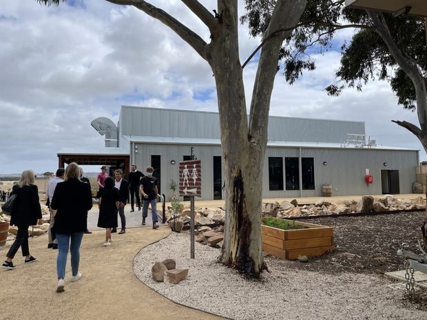 A low-rise contemporary building with a corrugated metal-clad upper facade and flat roof, viewed from a landscaped outdoor approach. Large dark-framed windows and glazed doors open onto a pale paved path, while native trees, gravel beds, rock landscaping and raised timber planter boxes surround the building. Several people are walking near the entrance; a broad water or landscape vista is visible beyond the site.