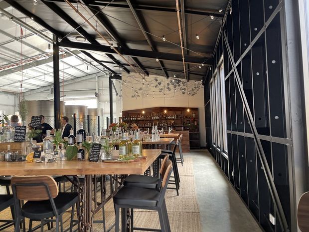 An expansive industrial-style bar and hospitality venue with exposed steel roof framing, visible services, pendant string lighting, timber communal tables, bar stools, and a fully stocked back bar. Large black-framed glazed or industrial partitioning lines the right side, while brewing or storage tanks and service areas are visible toward the rear. The space combines raw industrial construction with warm timber furniture and ambient lighting.