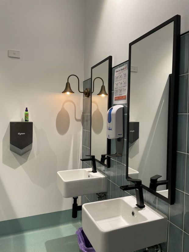 A compact contemporary bathroom or washroom with two wall-mounted white rectangular basins, matte black taps and exposed plumbing, large black-framed mirrors, teal-grey tiled wall sections, and matching teal flooring with a curved transition. Warm wall sconces illuminate the mirrors. A wall-mounted soap dispenser, hand dryer, and hygiene instruction sign indicate a commercial or public washroom setting.