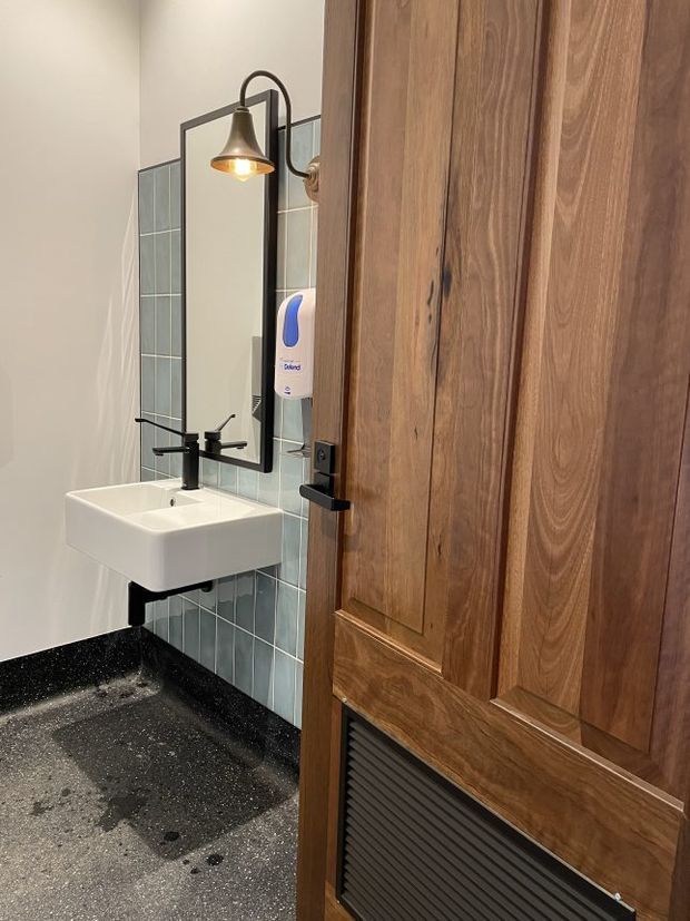 Compact contemporary bathroom viewed through a partially open timber door. The room features a wall-mounted white basin with a matte black tap, a black-framed rectangular mirror, a warm pendant wall light, pale blue vertical wall tiles, a black stone-like splashback and dark speckled flooring. The timber door has raised panels and a black louvred ventilation grille.