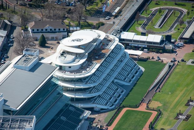 Aerial view of a large contemporary multi-storey sports or entertainment complex with sweeping curved white roof forms, extensive glass façades, and tiered structures. The building is surrounded by athletic fields, running tracks, paved circulation areas, and neighbouring urban facilities.