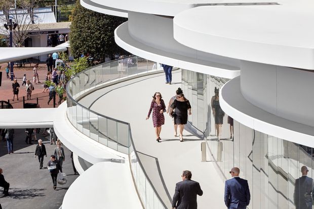 Exterior architectural view of a sculptural, multi-level white building with sweeping curved floor plates, rounded cantilevered balconies, extensive reflective glazing, and transparent glass balustrades. Pedestrians are walking along an elevated outdoor circulation terrace, with a busy public plaza visible below and behind.