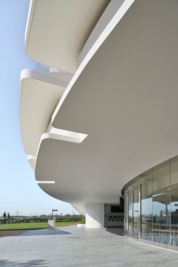 Modern institutional or commercial building exterior photographed beneath a dramatic cantilevered canopy. The structure features smooth white curved soffits and projecting slabs, large glazed entrance doors and curtain-wall glazing, and a broad grey paved forecourt. The setting appears open and landscaped, with a clean, sculptural contemporary architectural character.