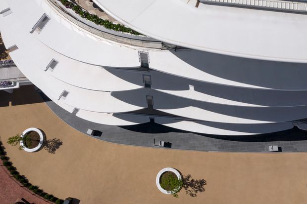 Aerial view of a large contemporary building with sweeping curved white roof forms, dark linear shadow bands, narrow roof openings, and a surrounding paved forecourt with circular planted features. The image emphasizes the building's sculptural roof geometry and expansive architectural composition.