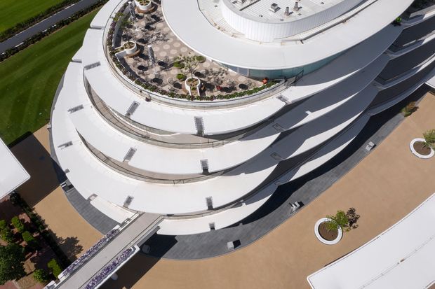 Aerial view of a large contemporary multi-level building with sweeping curved white roof terraces, layered balconies, extensive rooftop landscaping, and broad paved exterior areas with circular planted beds. The image focuses on the building's sculptural exterior form and roofscape.