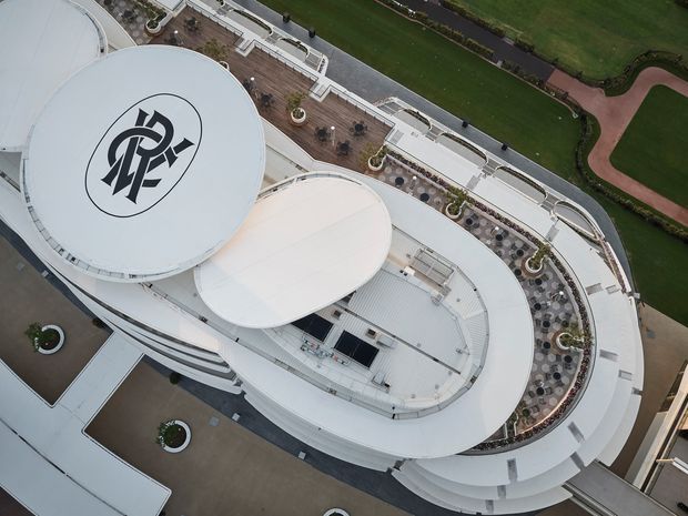 Aerial view of a large contemporary oval-shaped building with sweeping white curved roof forms, layered terraces, landscaped planters, and patterned outdoor seating areas. A prominent circular roof bears a bold black monogram logo. The surrounding grounds include manicured lawns and curved pedestrian paths.