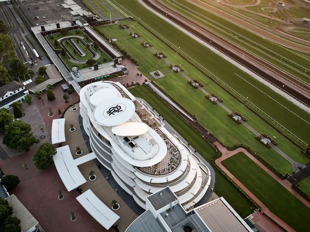 Aerial view of a large, sculptural white multi-level building situated beside an extensive horse-racing track. The building features curved layered terraces, rooftop areas, landscaped courtyards, and surrounding paved pedestrian spaces. Multiple racetrack lanes, manicured grass, and equestrian facilities dominate the adjacent grounds.