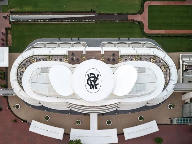 Aerial top-down view of a large oval-shaped contemporary building with a predominantly white architectural envelope, rounded roof forms, terraces, landscaped courtyards, and surrounding red and green recreational or campus surfaces. A prominent circular central roof bears a stylised black monogram.