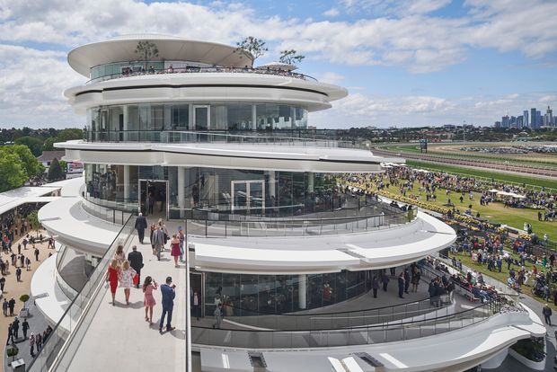 A large multi-level contemporary grandstand or hospitality building with sweeping curved white floor plates, extensive floor-to-ceiling glazing, rooftop terraces, and glass balustrades. Crowds gather around the building and on the adjacent racecourse grounds, with a distant urban skyline visible beyond.