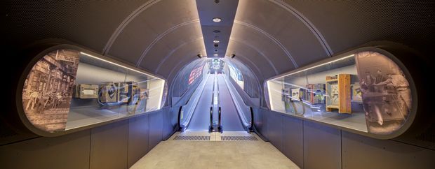 A futuristic enclosed interior with a central ascending escalator or moving walkway, curved ribbed ceiling, dark metallic wall panels, and illuminated recessed display vitrines on both sides. The displays contain historical or cultural imagery and objects, creating the atmosphere of a museum or exhibition passage. The symmetrical tunnel-like composition is cool-toned, refined, and dramatic.