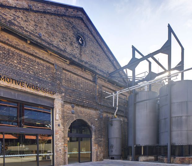 Exterior view of a converted historic brick industrial building, with arched openings, large glazed workshop frontage, faded painted lettering, and prominent steel structural framing and cylindrical industrial tanks. The scene combines heritage masonry with retained industrial infrastructure and contemporary additions.