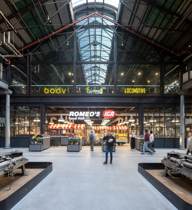 A large indoor market hall in a repurposed industrial building, with exposed steel trusses, dark metal framing, corrugated roofing, and a prominent central arched glass roof bringing in daylight. The ground floor is a food hall with a Romeo’s IGA grocery storefront, market stalls, shopping carts, benches, and pedestrians. Bright signage contrasts with the dark industrial structure.