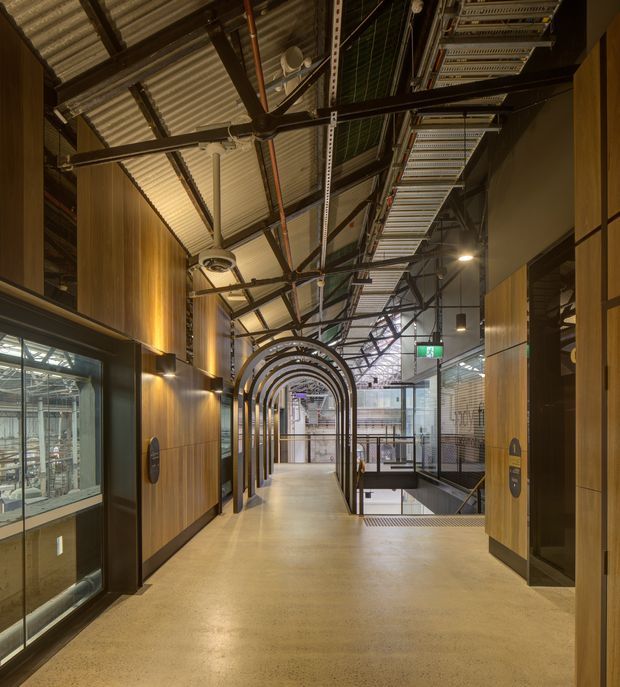 A contemporary commercial interior corridor with a polished concrete floor, warm timber wall panelling, glazed display or meeting-room fronts, and a sequence of dark metal arched frames leading toward a bright atrium. The exposed roof structure, services, cable trays, and corrugated metal ceiling create an industrial warehouse-like character, while integrated lighting and signage provide a refined workplace or public-building fit-out.