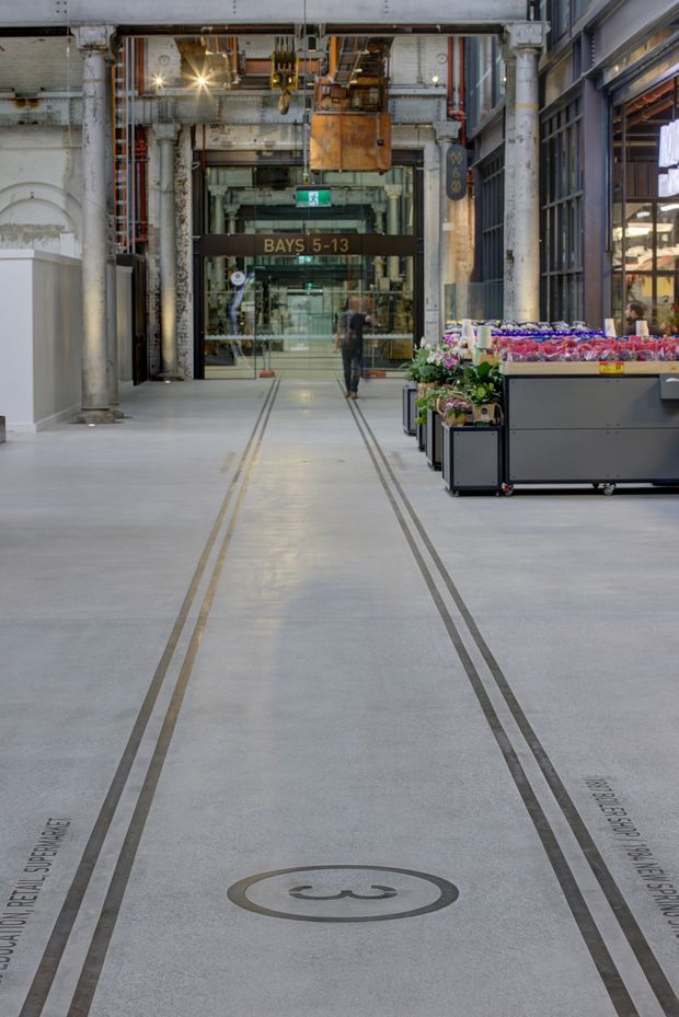 Interior view of a large adaptive-reuse commercial market hall or arcade. A broad polished concrete circulation floor is marked with parallel embedded metal rails and wayfinding text, leading toward a glazed entrance labelled “Bays 5–13.” The space retains substantial industrial fabric, including weathered structural columns, exposed overhead services, a suspended hoist or crane mechanism, and tall steel-framed glazing. A produce or flower market stall occupies the right side.