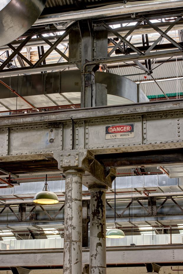 Vertical interior view of a large industrial building showing exposed riveted steel beams, columns, trusses, ductwork, corrugated roofing, suspended industrial pendant lights, and layered structural mezzanine or walkway elements. Weathered paint, raw metal, and visible services create a strong warehouse-conversion character. A prominent safety sign reads “DANGER LIVE WIRES.”