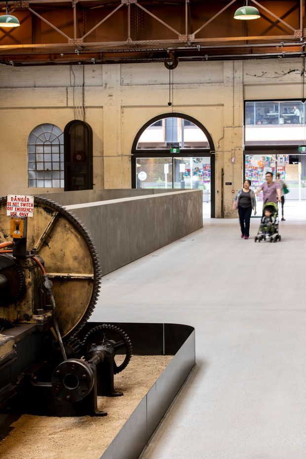 Interior view of a repurposed industrial building or museum, featuring preserved heavy machinery in the foreground, exposed steel roof trusses, aged painted masonry walls, arched windows and doorways, and a polished concrete circulation floor. Visitors are walking through the large open space, with urban buildings visible through the glazing.
