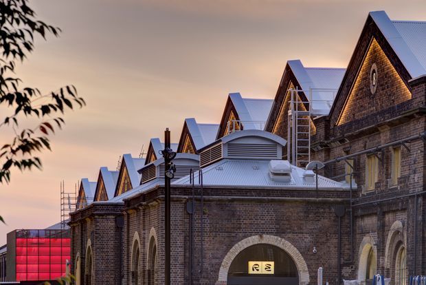 Exterior architectural photograph of a long historic brick building with repeated steep gabled roof forms, arched openings, masonry detailing, and modern rooftop additions. A vivid red glazed structure is visible at the far left, with warm dusk light and silhouetted foliage framing the scene.