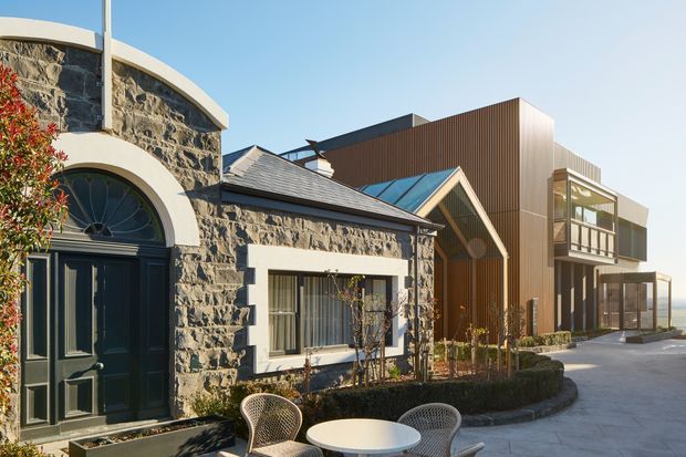 Exterior view of a contemporary residential development combining an older stone-clad building with arched openings and dark timber detailing, alongside a newer multi-level addition with vertical timber cladding, pitched roof elements, balconies, and large glazed areas. Paved forecourt and outdoor café-style seating are visible in the foreground.