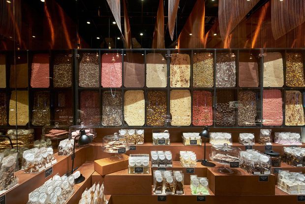 A warmly lit commercial retail display space featuring a wall of neatly arranged transparent-front compartments filled with colourful granular products, likely food or confectionery. Additional products are presented on stepped timber-toned display counters in the foreground. Dark reflective ceilings, timber wall accents, glass display panels, and focused spot lighting create a rich, boutique showroom atmosphere.