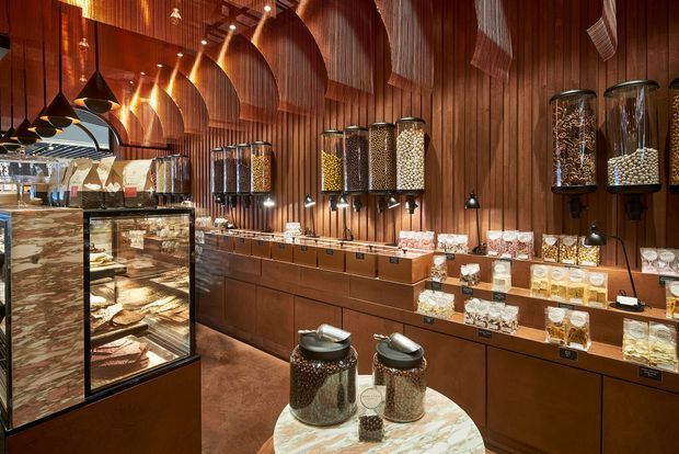A richly detailed specialty food retail interior, likely a confectionery or gourmet shop, with warm vertical timber wall panelling, curved ribbed ceiling elements, illuminated pendant and track lighting, glass display cases, wall-mounted bulk dispensers, and extensive built-in timber cabinetry and shelving. The palette is dominated by dark and mid-brown wood with warm amber lighting and marble display surfaces.