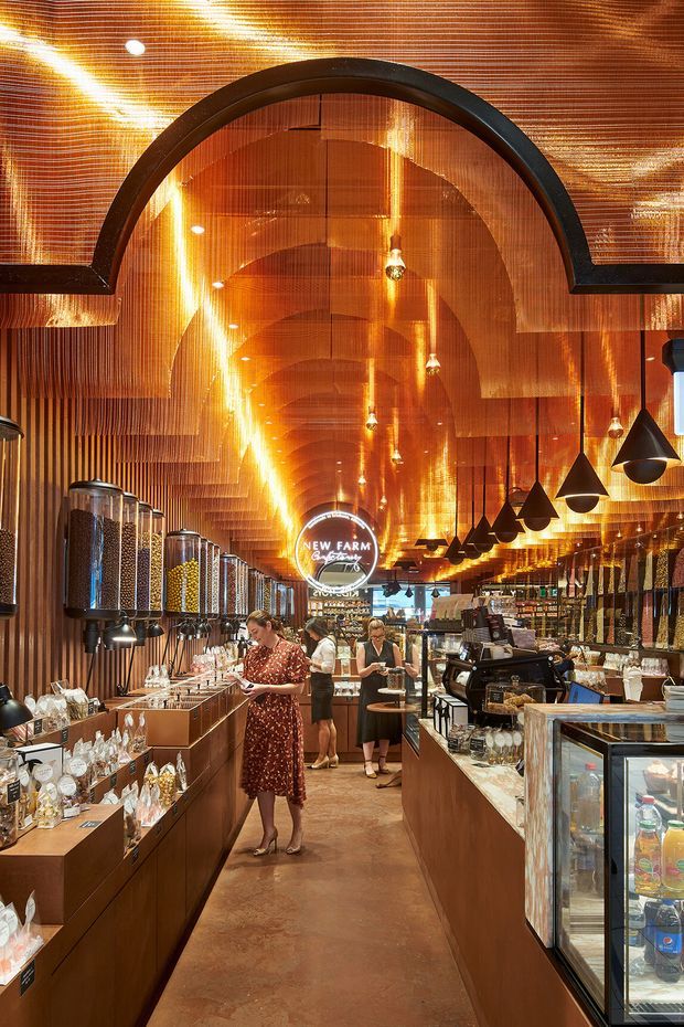 A richly detailed commercial retail interior, likely a specialty food or confectionery shop. The long narrow space has warm copper-toned ribbed wall and ceiling surfaces, repeated arched ceiling forms, dark pendant lights, timber-toned display counters, glass jars and dispensers, refrigerated display cases, and a central customer aisle. Several shoppers and staff are visible. The atmosphere is highly decorative, warm, and immersive, with a strong metallic amber palette.