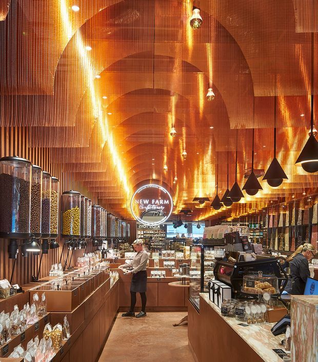A richly detailed commercial retail interior, likely a specialty food or coffee shop, with warm copper-toned curved ceiling panels and suspended chain-like ceiling installations. The space features rows of transparent dispensers, timber-toned counters and cabinetry, display shelving, hanging black pendant lights, a central service counter, and staff working behind the counters. The design is dramatic, immersive, and highly coordinated in amber, copper, and dark tones.