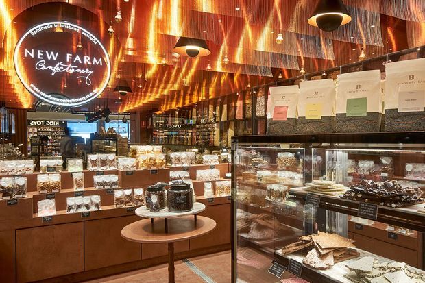 A richly detailed New Farm confectionery or chocolate shop interior, with extensive glass display cabinets and timber-toned counters showcasing packaged sweets, chocolates, and gift items. The space features a dramatic reflective metallic ceiling with warm amber lighting, dark framed glazing and shelving, pendant lights, and a prominent circular illuminated New Farm Chocolatier sign. The retail fit-out feels luxurious, warm, and highly curated.