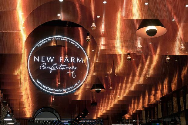 A dramatic confectionery shop interior featuring reflective copper-toned wall and ceiling cladding, a large illuminated circular New Farm Confectionery sign, and multiple pendant and recessed lights. The space has a dark, atmospheric, contemporary retail fit-out with curved architectural forms and extensive metallic surfaces.