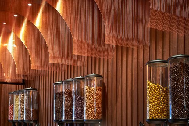 A warm, richly detailed retail display interior featuring rows of clear cylindrical dispensers filled with colourful dry goods or confectionery. The space is lined with vertical dark timber slats, while suspended sculptural timber screens or draped ribbed panels create a layered ceiling treatment. Small recessed spotlights provide focused illumination, producing a cozy and dramatic atmosphere.