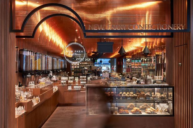 A richly detailed confectionery and café-style commercial interior with warm copper-toned ribbed wall and ceiling surfaces, dark metal arched detailing, illuminated display counters, glass pastry cases, shelving, pendant lights, and rows of bulk food dispensers. The space has a boutique retail feel with visible confectionery merchandise and signage reading New Farm Confectionery.