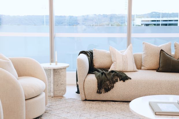 Bright, softly styled living room with a curved cream boucle armchair and matching sofa, layered neutral cushions, a dark fringed throw, a small marble side table, and a round white coffee table. Floor-to-ceiling windows provide expansive views beyond the room, while a pale patterned rug and light finishes create an airy, refined atmosphere.