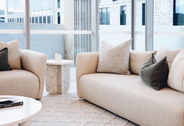 A bright, refined contemporary living room featuring a large beige upholstered sofa and armchair, neutral textured cushions, a round white coffee table, a small stone side table, and a pale patterned rug. Floor-to-ceiling glazing provides views toward a light-toned urban building exterior.