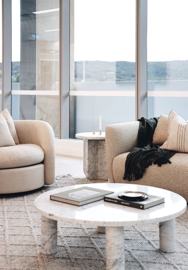 A bright, refined living room with two rounded upholstered sofas and armchairs arranged around a large round marble coffee table. Floor-to-ceiling glazing provides expansive views toward wooded hills, while a smaller marble side table, neutral textured rug, cushions, and a dark throw create a soft, contemporary setting.