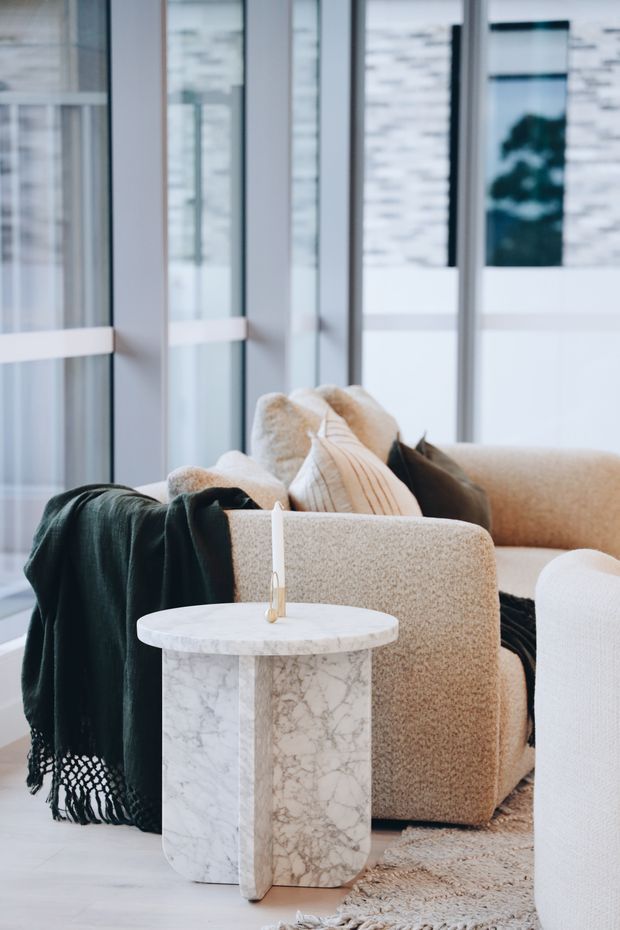 A softly styled contemporary living room vignette with rounded cream boucle armchairs and sofa, layered neutral cushions, a dark green throw, and a cylindrical white marble side table holding a slender candle. Tall floor-to-ceiling glazed panels create a bright, airy backdrop, with pale flooring and a textured neutral rug completing the scene.