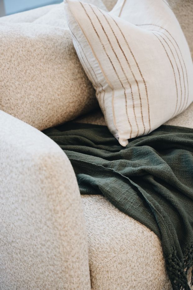 Close-up detail of a softly upholstered beige sofa or bed with layered cream and beige cushions, including a striped decorative pillow, and a dark olive-green textured throw draped across the seating. The image focuses on tactile fabrics and a relaxed, neutral interior styling.