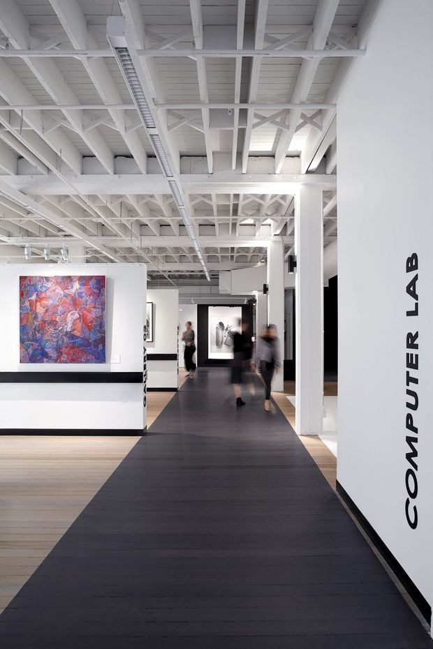 A bright contemporary computer lab or gallery-like commercial interior with white walls, exposed white-painted structural roof trusses and services, black display plinths, and a dark timber walkway leading through the space. Large colourful artworks and framed displays line the walls, with blurred visitors moving through the corridor. The word “COMPUTER LAB” is displayed vertically on the foreground wall.