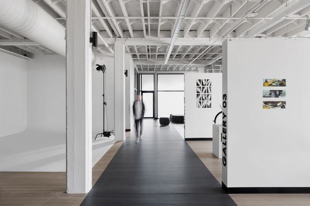 Bright, minimalist gallery interior with white walls, exposed white-painted structural ceiling beams and services, tall black-framed glazing, and a contrasting dark linear floor strip through the circulation zone. Artwork and display pieces are visible, with gallery signage on a central partition and sparse contemporary furnishings in the background.