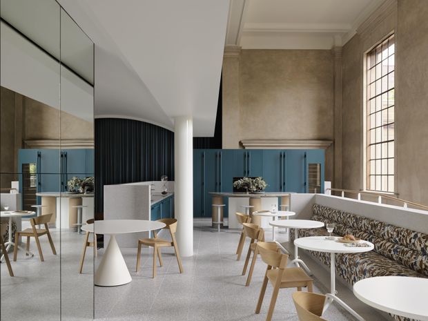 A refined contemporary hospitality or café interior in a tall, heritage-like room. The space features blue fixed cabinetry and a service counter, a long patterned upholstered banquette, white round tables, light timber chairs, a curved black fluted screen, a large gridded window, mirrored wall panels, pale terrazzo flooring, and ornamental ceiling cornices.