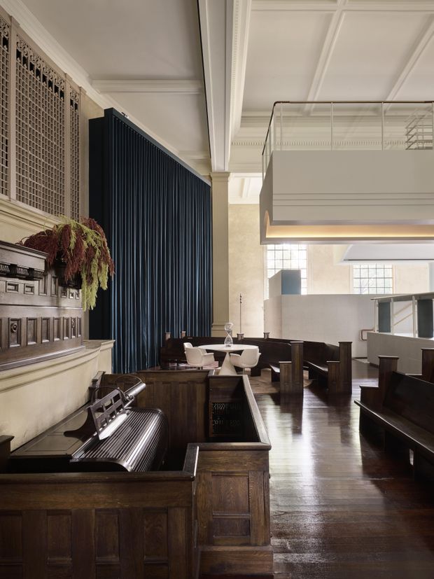 A grand, heritage-style interior with dark timber pews and panelled joinery, a polished timber floor, ornate decorative wall grilles, classical mouldings and a high coffered ceiling. A large suspended mezzanine or balcony with a translucent balustrade overlooks the seating area. The space appears to be a church or chapel adapted as a contemporary hospitality or commercial venue, with modern tables and chairs among the original architectural features.