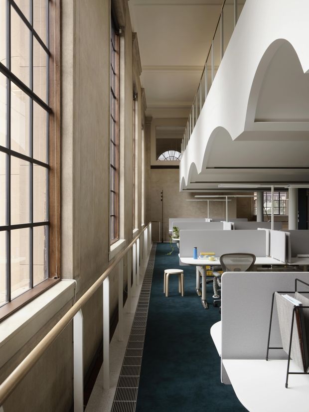 A refined open-plan office interior in a tall, historic-looking space, viewed along a corridor of workstations beside large multi-pane industrial-style windows. The room features deep blue carpet, pale grey and white desk partitions, white desks, contemporary office chairs, and prominent curved white architectural ceiling or canopy elements. Tall columns, decorative arch details, clerestory glazing, and warm plaster-toned walls combine heritage architectural character with modern workplace fit-out.