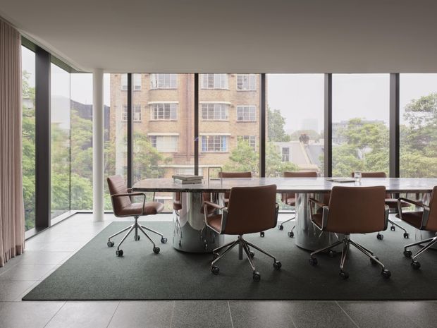 A bright contemporary meeting room with a long conference table and multiple brown swivel office chairs. Floor-to-ceiling glazing wraps around the room, providing views of surrounding greenery and neighbouring buildings. A large dark area rug sits over a tiled floor, with curtains along the left wall and a clean, professional atmosphere.