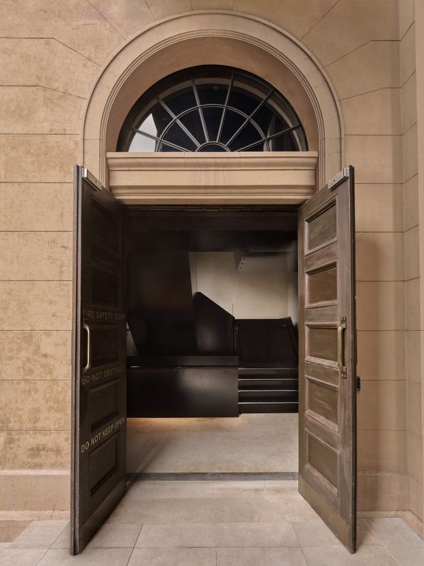 A formal architectural entrance with heavy dark timber double doors standing open beneath a large arched fanlight window. The surrounding facade is clad in warm beige stone with horizontal joints and classical moulded arch detailing. Inside is a dark, compact stair or service lobby with black finishes and a short flight of steps.