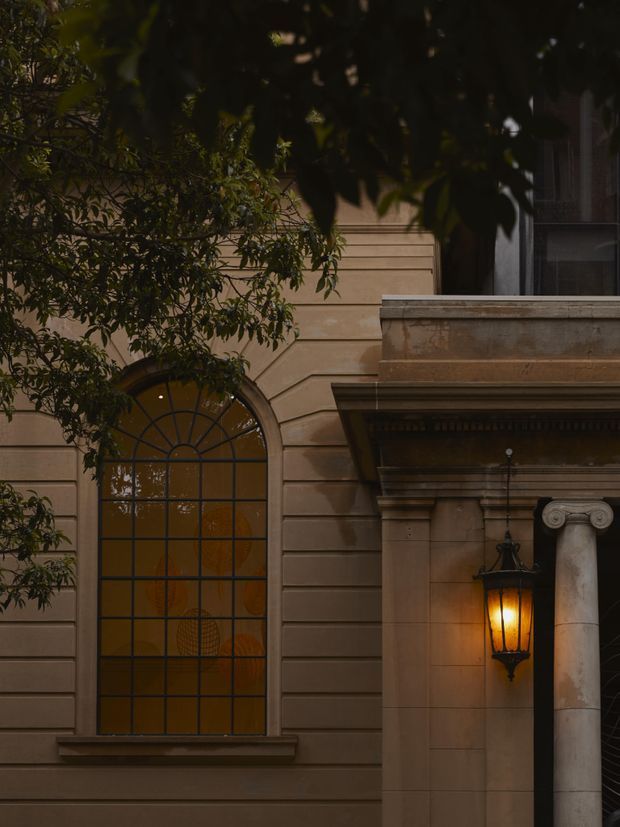 Close-up exterior architectural detail of a stately, traditional facade. The image shows warm brown horizontal rendered or stone banding, a large arched leaded-glass window, substantial classical cornice and entablature detailing, a pale stone column with Ionic-style capital, and a glowing hanging lantern. Dark foliage partially frames the upper and left portions of the facade, creating a moody, refined atmosphere.