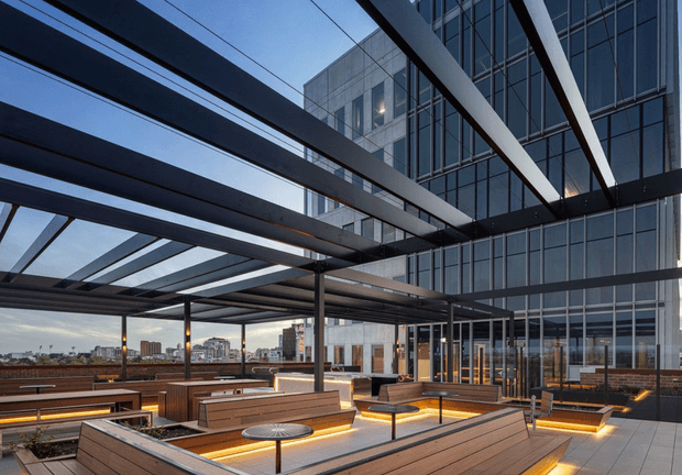 A contemporary rooftop terrace or elevated outdoor lounge beside a tall glazed commercial building. The space features extensive steel pergola beams, timber-clad built-in bench seating, small round tables, integrated warm LED lighting, and large areas of glass and metal framing. A dense urban skyline is visible beyond the terrace.